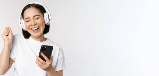 Schöne moderne asiatische Mädchen, Musik über Kopfhörer hören, Handy in der Hand, Smartphone-App, vor weißem Hintergrund stehen - Foto, Bild