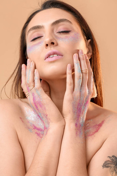 Mooie jonge vrouw met glitters op haar gezicht en lichaam tegen kleur achtergrond - Foto, afbeelding