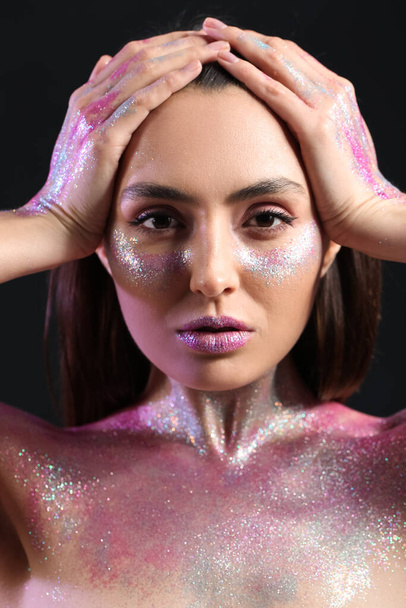 Beautiful young woman with glitters on her face and body against dark background - Photo, Image