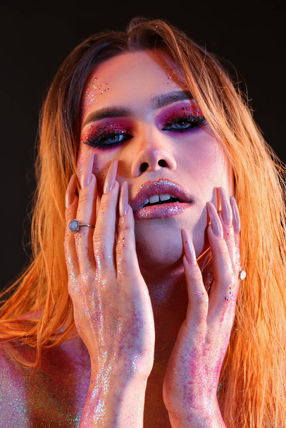 Modieuze jonge transgender vrouw met glitters op het lichaam tegen donkere achtergrond - Foto, afbeelding