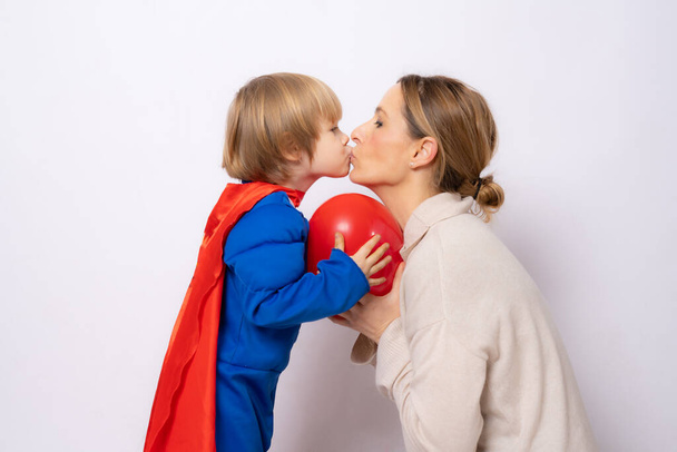 Child boy dressed in superman outfit kissing his mother isolated over white background. Motherhood concept. - Valokuva, kuva