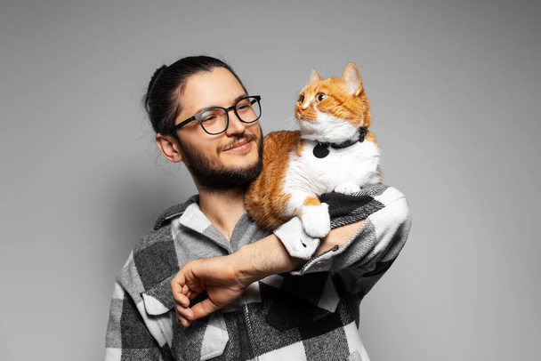 Studio ritratto di giovane uomo sorridente con gatto rosso sulle braccia. Fondo grigio. - Foto, immagini