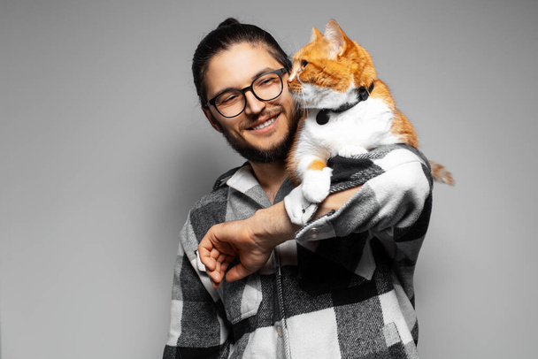 Felice giovane uomo e gatto rosso-bianco su sfondo grigio. - Foto, immagini