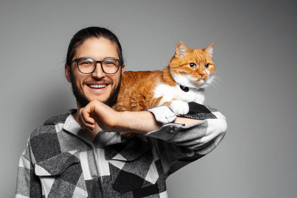 Studio portrait of young happy man with red cat on grey background. - Photo, Image