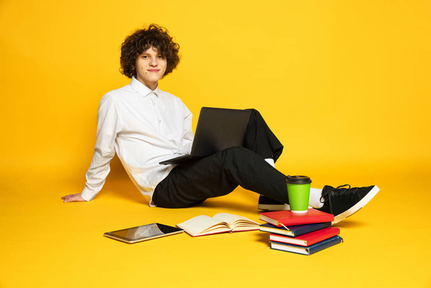 Portrait of young boy, student sitting on floor and studying isolated over yellow studio background - Foto, afbeelding