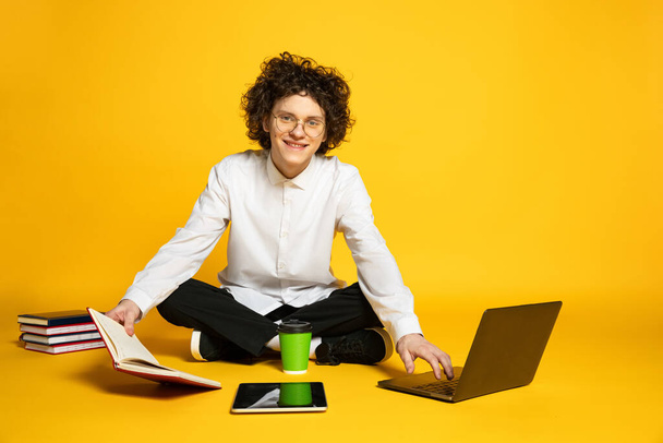 Retrato del joven sonriente, estudiante mirando a la cámara, posando alrededor de la computadora portátil y libros aislados sobre fondo amarillo del estudio - Foto, imagen
