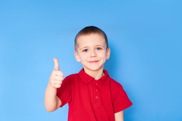 charming little smiling boy in a red shirt shows thumbs up, on a blue background. - Фото, зображення