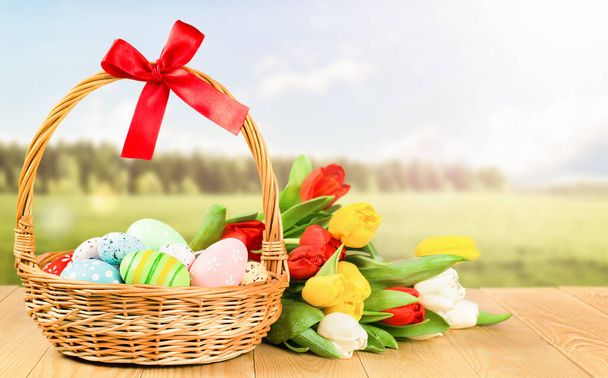 cesto di vimini con uova di Pasqua dipinte e un mazzo di tulipani su un tavolo di legno sullo sfondo della natura - Foto, immagini