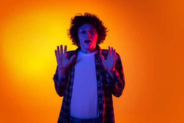 Half-length portrait of young curly man in checkered shirt showing emotion of shock isolated over orange background in neon light - Foto, afbeelding