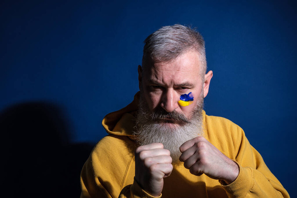 Felnőtt szakállas férfi öltözött sárga kapucnis felhívja ukrán zászló az arcára, tiltakozás a háború ellen - Fotó, kép