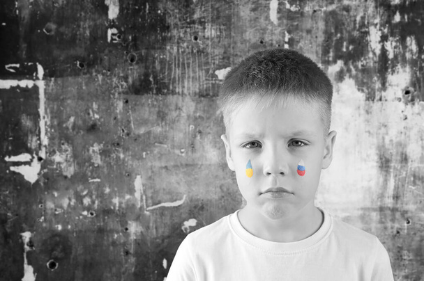 少年は、ウクライナとロシアの旗の色で描かれた彼の顔に涙の滴と暗い、ショットの壁の背景に立っています。戦争はない。子供は平和を望む. - 写真・画像