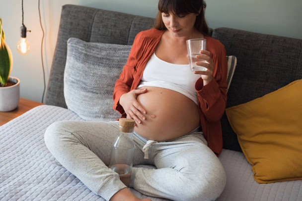 Pregnant woman holding glass of water relaxing in bed at home - Фото, изображение