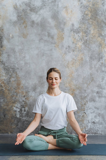 Happy girl in a tracksuit, sits on the floor in the lotus pose. A lady meditates,crossed fingers in Yoga Mudra. Assanas for Meditation and Relax. High quality photo - 写真・画像