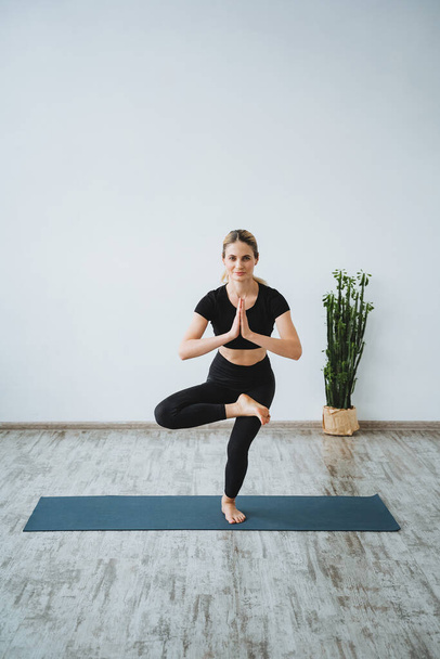 A fit young girl does yoga in the studio.Perform Tree pose. Assana Vrishkasana.Meditation and relaxation after yoga. spacious yoga studio. High quality photo - Foto, afbeelding