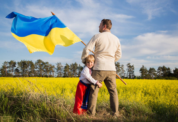 apa és fia együtt állnak Ukrajna sárga és kék zászlajával a virágzó repcemagmező hátterében. Hazafias nevelés. A szabadság és függetlenség nemzeti szimbóluma. Ukrajna támogatása - Fotó, kép