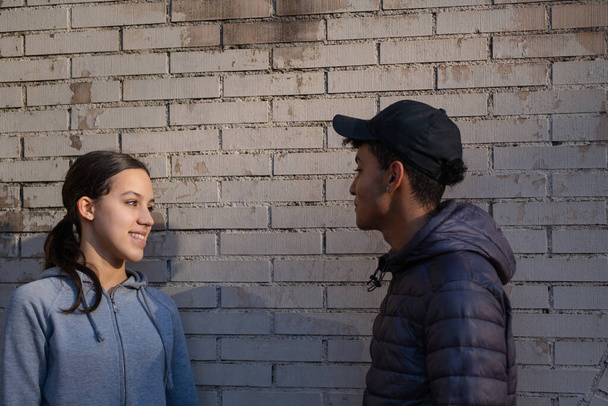 Young couple talking and smiling. A couple standing with a brick wall behind. The guy looks at the girl with tenderness. Love, relationship, commitment, romantic. - 写真・画像