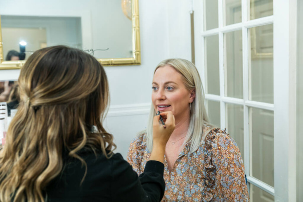 Kosmetikerin schminkt eine blonde Frau im Schminkstuhl mit Lippenstift - Foto, Bild