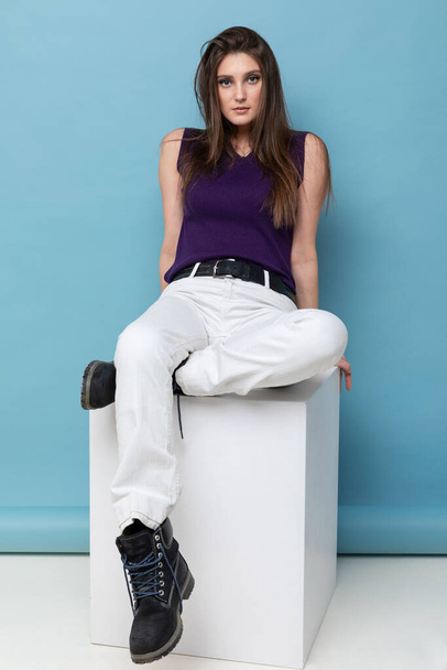 Fashion portrait of a young woman in a tank top and light pants on a blue background - Fotó, kép