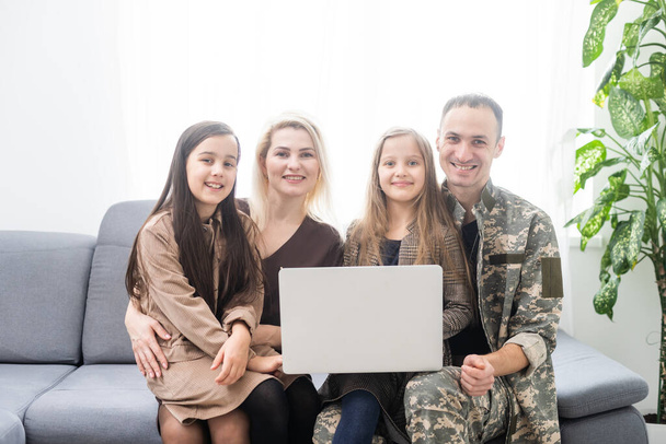 Ευτυχισμένος στρατιωτικός που χαμογελάει και αγκαλιάζει την οικογένειά του ενώ χρησιμοποιεί φορητό υπολογιστή σε εσωτερικούς χώρους. - Φωτογραφία, εικόνα