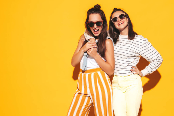 Two young beautiful smiling blond hipster female in trendy summer clothes. Sexy carefree women posing near yellow wall in studio. Positive models having fun. Cheerful and happy. In sunglasses - 写真・画像