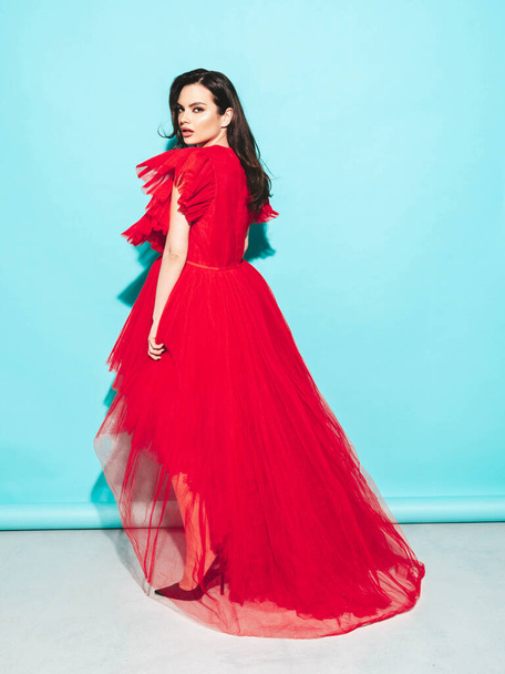 High fashion portrait of young beautiful brunette woman wearing nice trendy red evening fluffy dress. Sexy fashion model posing in studio. Fashionable female isolated on blue - Фото, зображення