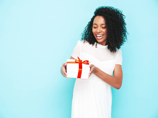 Beautiful black woman with afro curls hairstyle. Smiling model dressed in white summer dress. Sexy carefree female posing near blue wall in studio. Tanned and cheerful. Holding gift box. Isolated - Фото, зображення