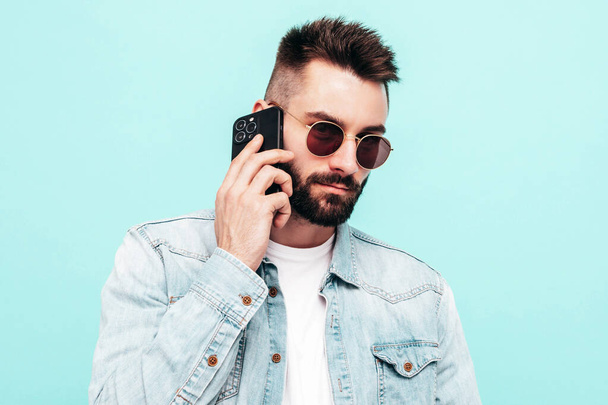 Handsome confident model.Sexy stylish man talking at smartphone. Fashion hipster male posing near blue wall in studio. Holding phone. With cellphone. In sunglasses. Isolated - Photo, Image