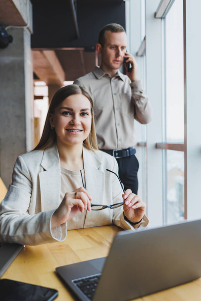 テーブルに座って眼鏡をかけたラップトップの後ろにいる若いビジネス女性は、現代のオフィスで同僚と企業のビジネスミーティングをしています。ビジネスキャリアの概念。フリースペース、選択的フォーカス - 写真・画像