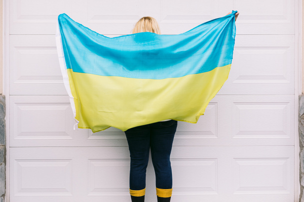 Ουκρανική ξανθιά-μαλλιά ώριμη γυναίκα από πίσω με κυματίζει μπλε και κίτρινη ουκρανική σημαία σε λευκό φόντο στο δρόμο. Πόλεμος στην Ουκρανία, εισβολή, διαμαρτυρία, πατριωτισμός και καμία πολεμική έννοια. - Φωτογραφία, εικόνα