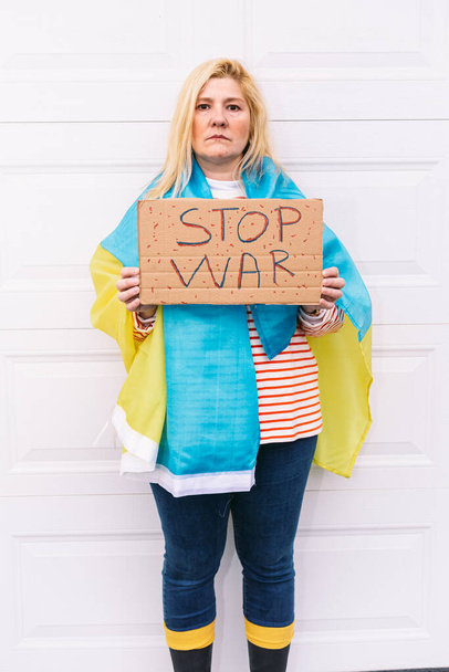 Donna ucraina con i capelli biondi e un gesto serio, con una bandiera ucraina blu e gialla con un cartello che recita: 'Stop War', per strada. Concetto Ucraina guerra, invasione, protesta, patriottismo. - Foto, immagini