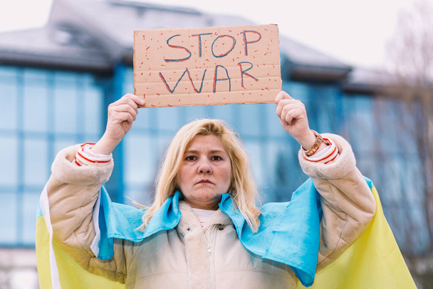 Ukraińska kobieta z blond włosami i poważnym gestem, z niebiesko-żółtą ukraińską flagą ze znakiem: "Stop wojnie", na ulicy, protestując przeciwko wojnie Ukraina-Rosja. - Zdjęcie, obraz