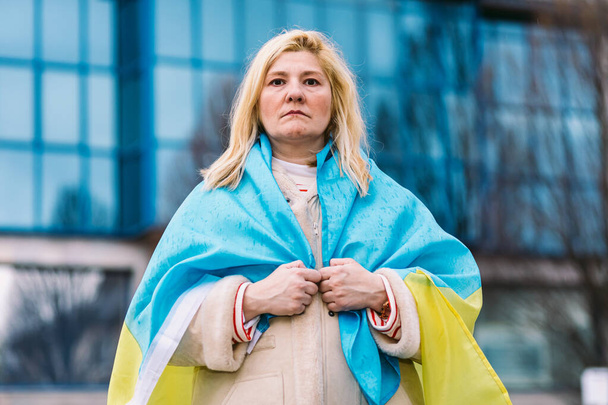 Donna ucraina con i capelli biondi e un gesto serio, con una bandiera ucraina blu e gialla, in strada, protestando contro la guerra Ucraina-Russia. - Foto, immagini