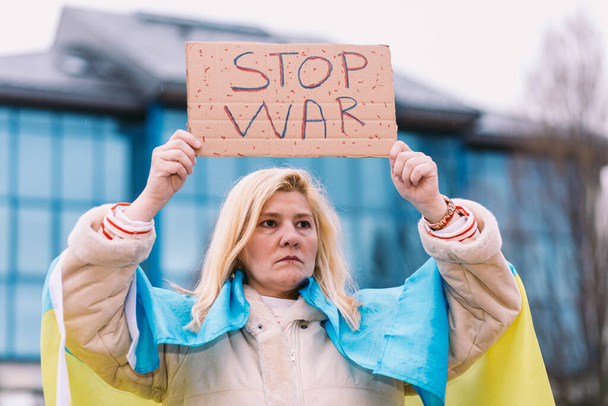 Oekraïense vrouw met blond haar en serieus gebaar, met een blauwe en gele Oekraïense vlag met een bord waarop staat: "Stop oorlog", op straat, protesterend tegen de oorlog tussen Oekraïne en Rusland. - Foto, afbeelding