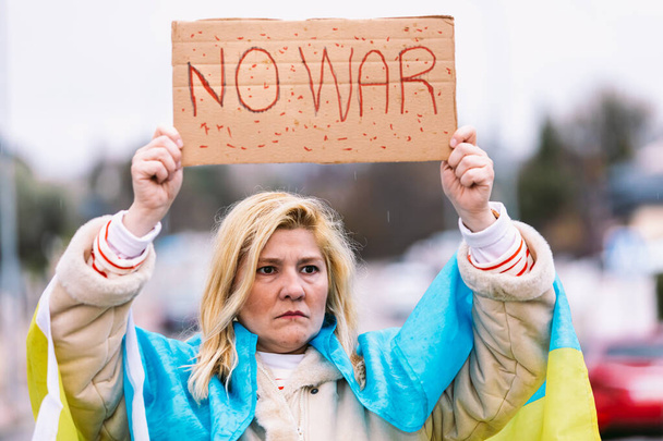 Ukraińska kobieta z blond włosami i poważnym gestem, z niebiesko-żółtą ukraińską flagą ze znakiem "No War", na ulicy, protestująca przeciwko wojnie ukraińsko-rosyjskiej. - Zdjęcie, obraz