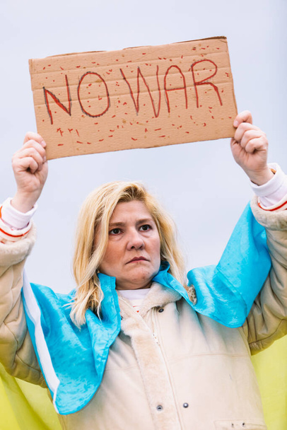 Ukrainische Frau mit blonden Haaren und ernster Geste, mit einer blau-gelben ukrainischen Flagge mit einem Schild mit der Aufschrift "No War", auf der Straße, um gegen den Krieg zwischen der Ukraine und Russland zu protestieren. - Foto, Bild