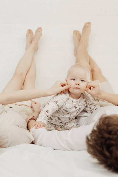 Young couple in white clothes lying with legs raised straight up high on bed tweaking cheeks of plump blue-eyed baby.  - Foto, Bild
