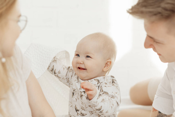 Portret pięknego, uśmiechniętego, niebieskookiego, pulchnego niemowlęcia siedzącego między rodzicami z białym bawełnianym kocem. - Zdjęcie, obraz