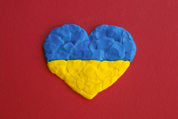 Ukrajinská vlajka. Srdce tvar modré a žluté plasticin modelování hlíny na červeném kaštanové textury pozadí. Plastikový prst texturované pozadí. Horní pohled, detailní záběr. - Fotografie, Obrázek