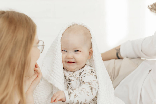 Gülümseyen mutlu mavi gözlü tombul bebeğin portresi beyaz battaniyeyle örtülmüş yatak örtüsünde oturuyor.. - Fotoğraf, Görsel