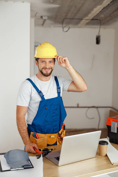 Lächelnder Handwerker in Uniform mit Werkzeuggürtel und Hut, der bei Renovierungsarbeiten drinnen in die Kamera schaut - Foto, Bild