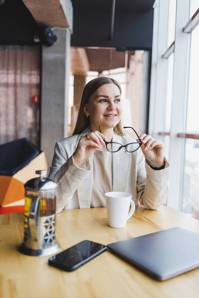 Eine junge Managerin in heller Jacke und Brille, ein Mädchen mit einem Lächeln im Gesicht, sitzt an einem Holztisch mit Laptop und Telefon. Fernbedienung - Foto, Bild