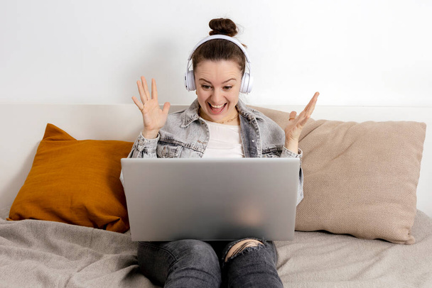 Молодая белая женщина в наушниках сидит на кровати с ноутбуком. Девушка использует блокнот для серфинга в Интернете, читает новости, смотрит кино, учится. Удивлённые, позитивные эмоции. Выиграл приз онлайн. - Фото, изображение