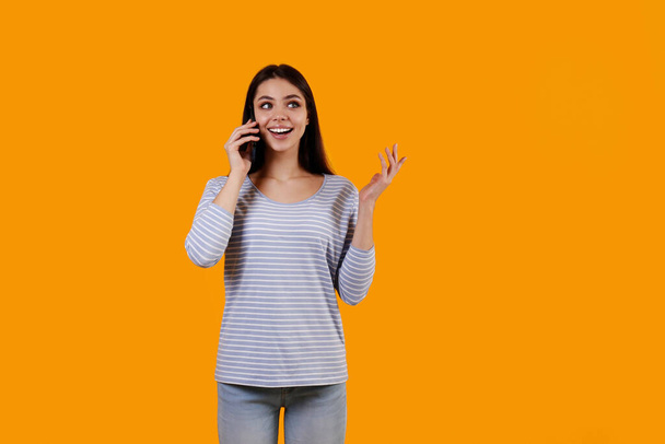 Portrait of young brunette woman wearing striped shirt talking on the phone and smiling. Joyful female model having fun phone call. Isolated yellow background, copy space, close up. - Fotografie, Obrázek