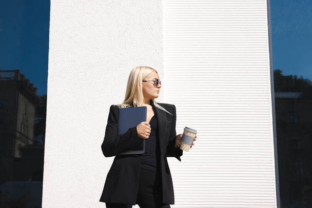 Una mujer joven en un traje elegante con un ordenador portátil y una taza de café posa sobre el fondo de un centro de negocios - Foto, Imagen
