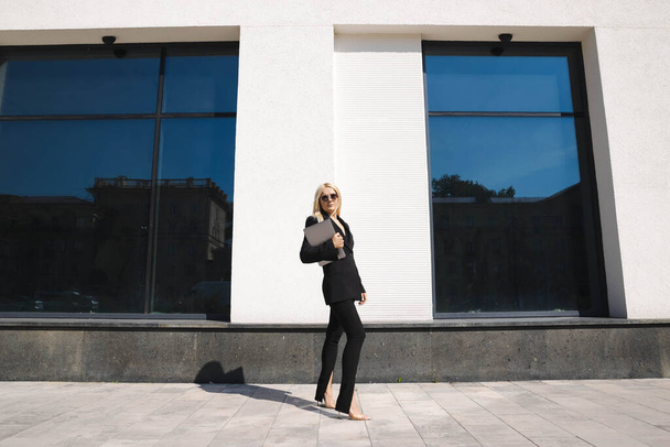 Uma jovem em um terno elegante com um laptop posa contra o pano de fundo de um centro de negócios - Foto, Imagem