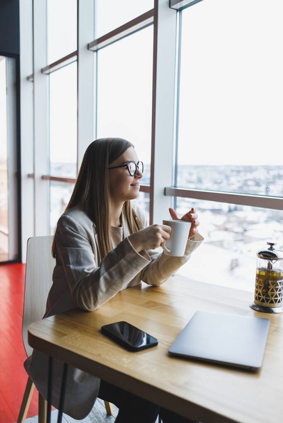 Een moderne vrouw van Slavisch uiterlijk, een manager in een lichte jas en bril, een meisje met een glimlach op haar gezicht, zit aan een tafel in een café en drinkt thee. Afstandswerk - Foto, afbeelding