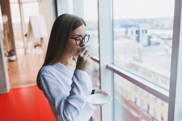 Mladá žena v brýlích evropského vzhledu, v bílé košili popíjí kávu a dívá se z okna - Fotografie, Obrázek