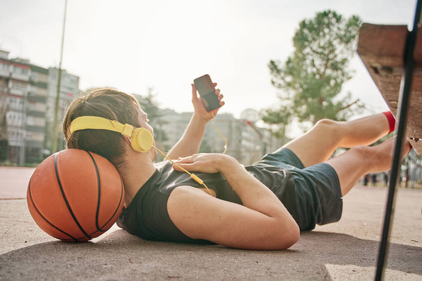basketball player lying down after training. leaning on basketball. making a video call with smart phone with yellow earphones. - Foto, Imagen