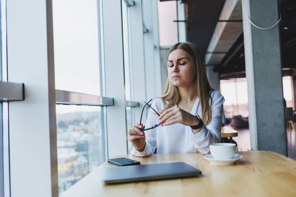 Een jonge vrouwelijke manager zet een bril op en gaat aan het werk, ze zit aan haar bureau in een wit shirt - Foto, afbeelding