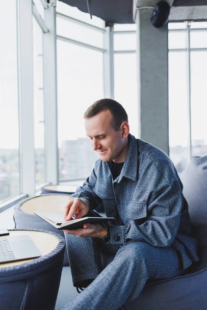 Um jovem estudante do sexo masculino em um terno casual descansando em um pufe confortável enquanto estuda faz anotações em um caderno, um freelancer atraente em um terno funciona remotamente - Foto, Imagem